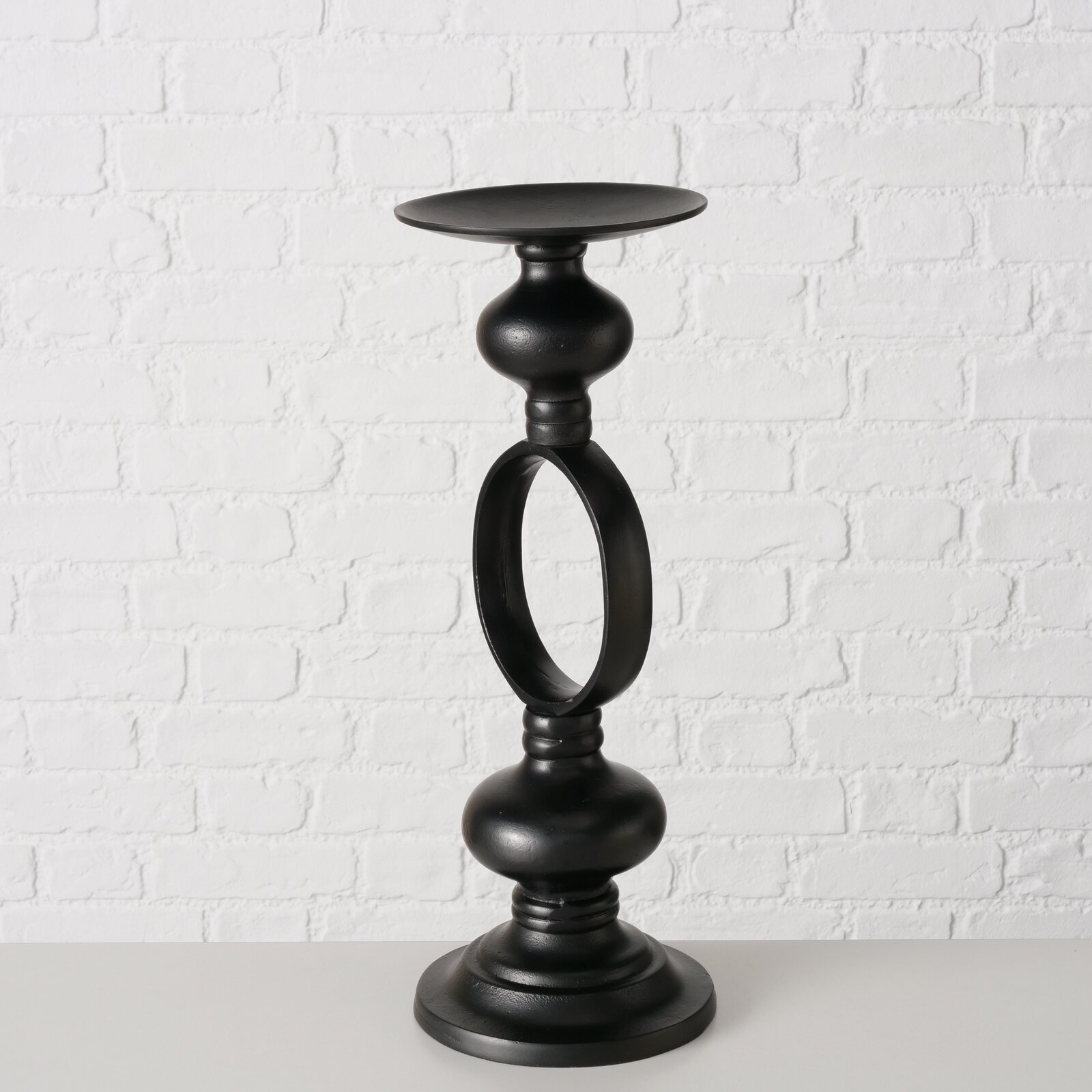 Schwarzer Kerzenleuchter Plokky aus Metall, frontal vor einer weißen Ziegelwand fotografiert.