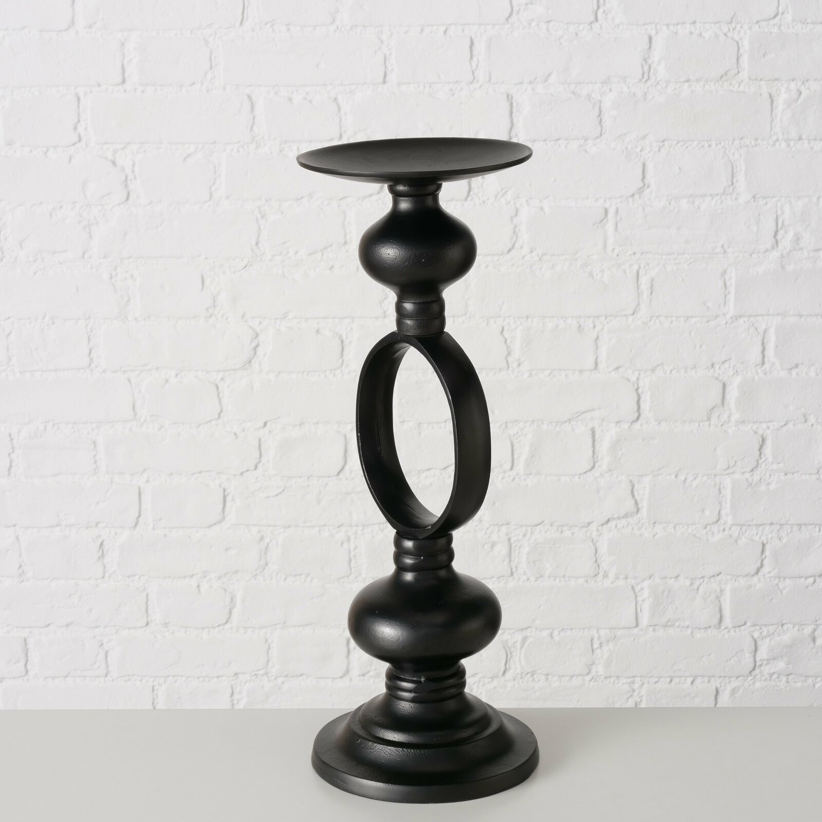 Schwarzer Kerzenleuchter Plokky aus Metall, frontal vor einer weißen Ziegelwand fotografiert.