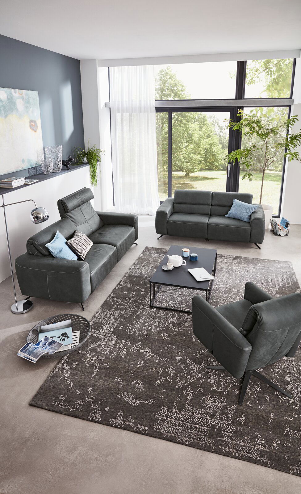 Modernes Wohnzimmer mit zwei grauen Sofas und einem Sessel, Blick von oben. Große Fenster mit Blick ins Grüne, stilvolle Dekoration und ein eleganter Teppich.