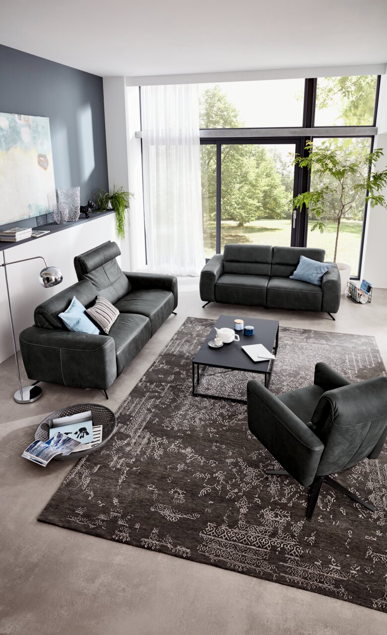 Modernes Wohnzimmer mit zwei grauen Sofas und einem Sessel, Blick von oben. Große Fenster mit Blick ins Grüne, stilvolle Dekoration und ein eleganter Teppich.