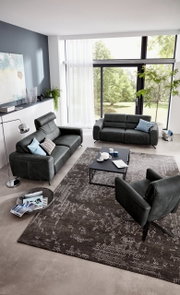 Modernes Wohnzimmer mit zwei grauen Sofas und einem Sessel, Blick von oben. Große Fenster mit Blick ins Grüne, stilvolle Dekoration und ein eleganter Teppich.