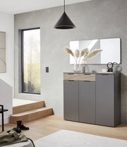 Moderne graue Kommode/Schuhschrank mit geriffelter Holzfront und schwarzen Griffen im Flur unter einem Wandspiegel, perspektivisch in leichter Seitenansicht von vorn rechts