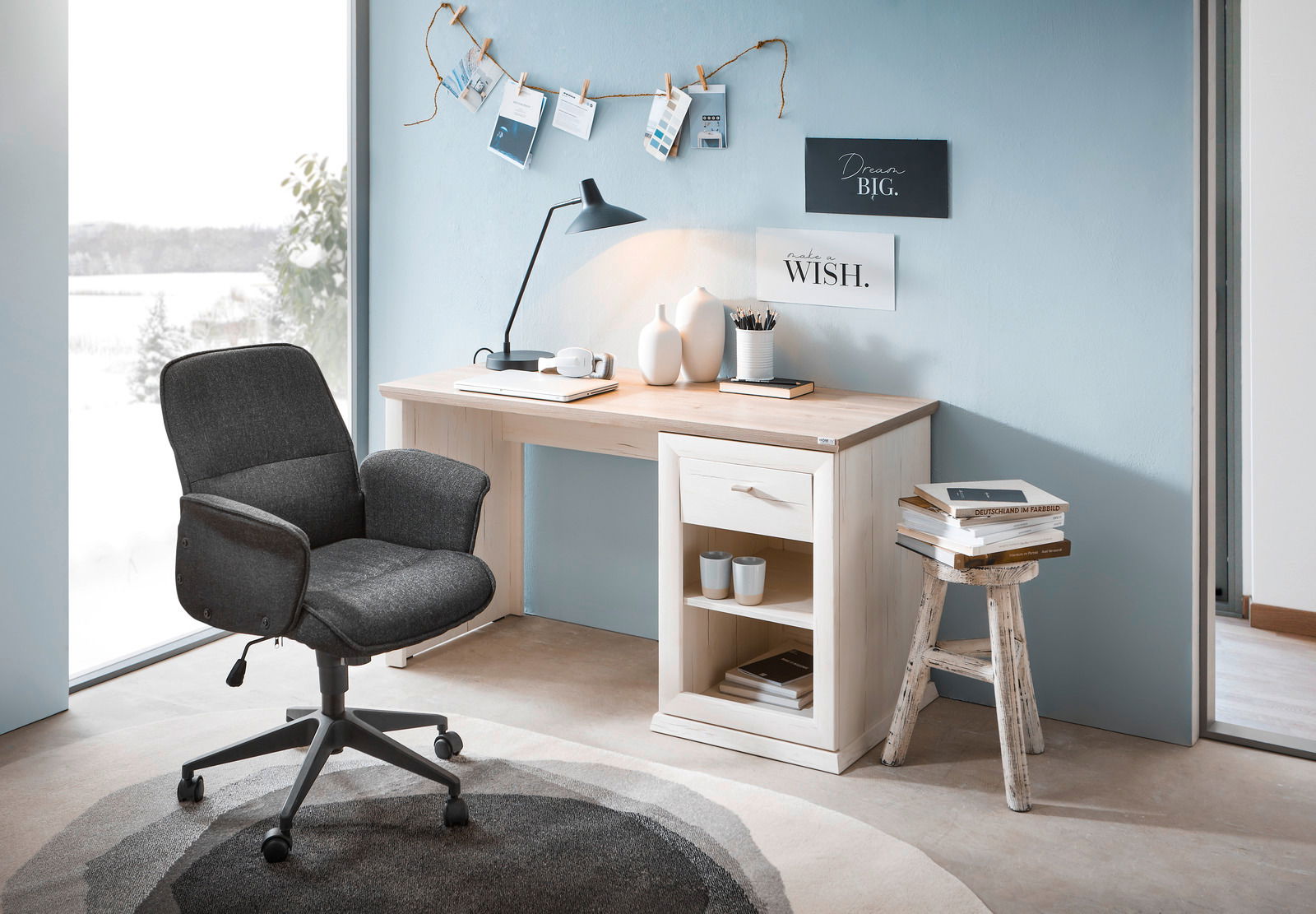 Moderner Schreibtisch in heller Holzoptik mit Schublade und Regal, seitliche Perspektive, dekoriert mit Lampe, Vasen und Büchern, neben einem großen Fenster mit Blick auf verschneite Landschaft.