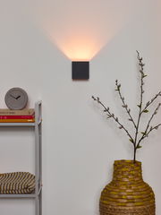 Moderne Wandleuchte XIO in dunklem Design, an der Wand montiert, mit warmem Licht nach oben abstrahlend, im Wohnzimmer mit dekorativen Elementen im Vordergrund.