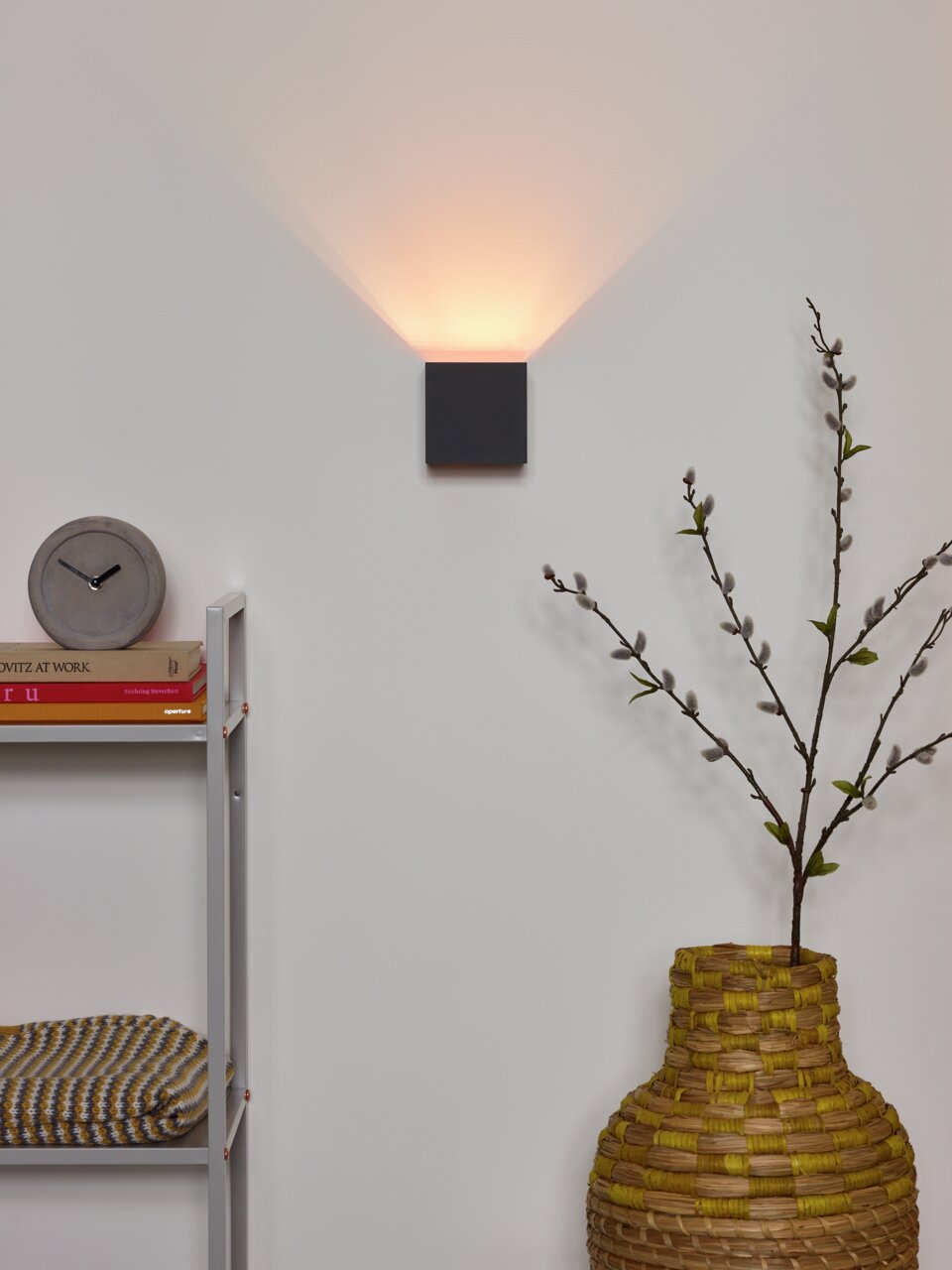 Moderne Wandleuchte XIO in dunklem Design, an der Wand montiert, mit warmem Licht nach oben abstrahlend, im Wohnzimmer mit dekorativen Elementen im Vordergrund.