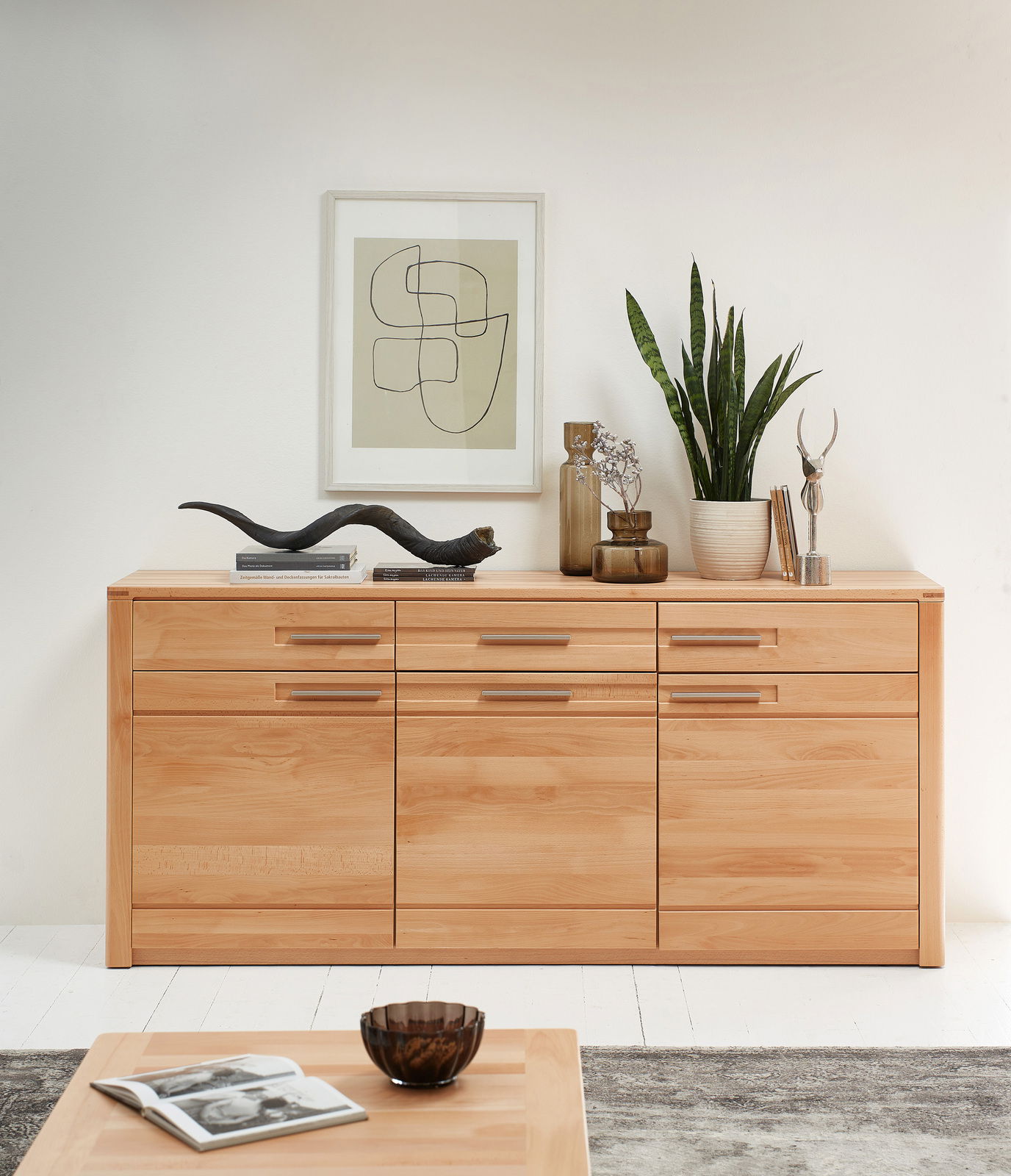 Holz-Sideboard mit drei Schubladen und drei Türen, dekoriert mit Pflanzen und Kunstobjekten, frontal fotografiert.
