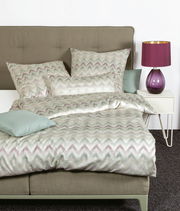 Bett mit Mako-Satin Bettwäsche in Zickzack-Muster, Perspektive von vorne, mit lila Lampe auf Nachttisch.