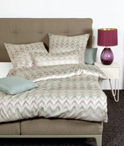 Bett mit Mako-Satin Bettwäsche in Zickzack-Muster, Perspektive von vorne, mit lila Lampe auf Nachttisch.