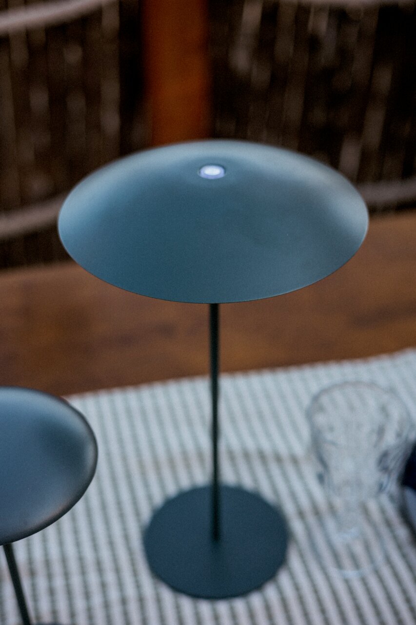 Moderne, ronde staande lamp in mat donkergrijs met een slanke voet en een platte, schijfvormige kap, van bovenaf licht schuin gefotografeerd.