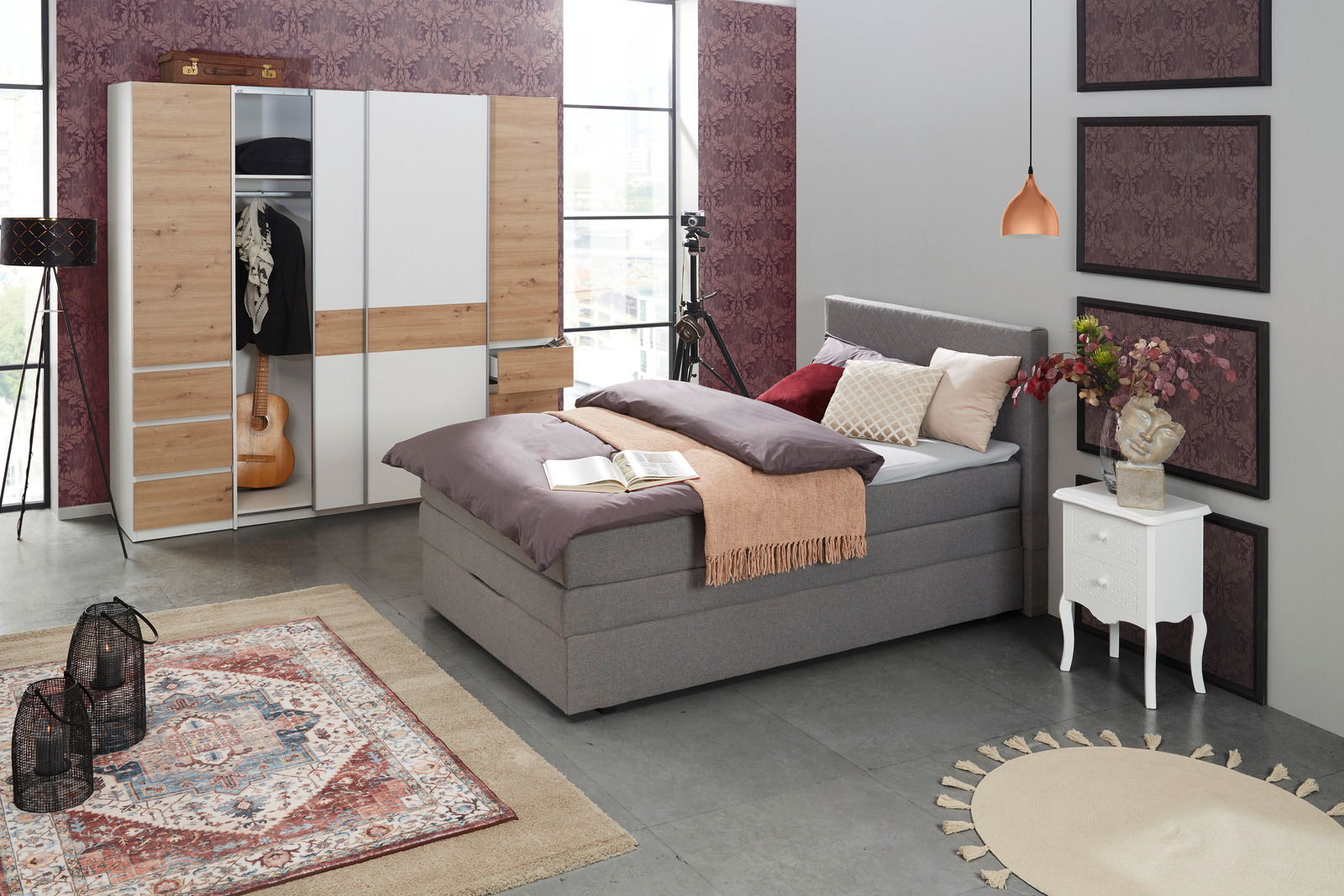 Schlafzimmer mit einem modernen Dreh- und Schwebetürenschrank in Holzoptik und Weiß, geöffnet, mit Kleidung und Gitarre. Perspektive von der Seite.