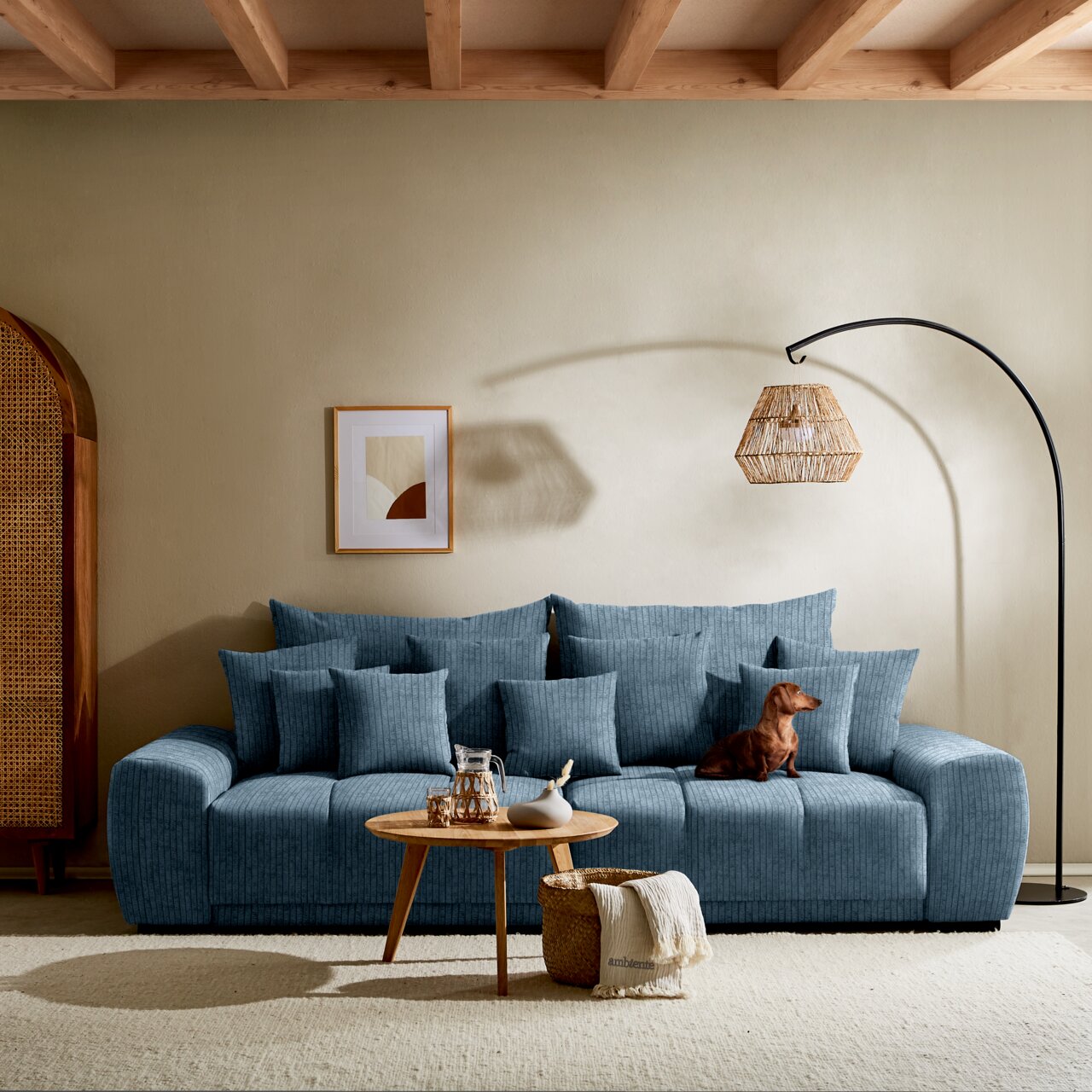 Großes, gemütliches Big-Sofa in Blau mit mehreren Kissen, aus der Frontalperspektive fotografiert. Ein kleiner Hund sitzt auf dem Sofa. Im Vordergrund steht ein runder Holztisch mit Dekoration.
