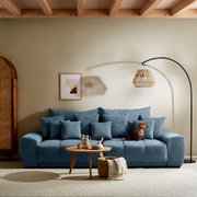 Großes, gemütliches Big-Sofa in Blau mit mehreren Kissen, aus der Frontalperspektive fotografiert. Ein kleiner Hund sitzt auf dem Sofa. Im Vordergrund steht ein runder Holztisch mit Dekoration.