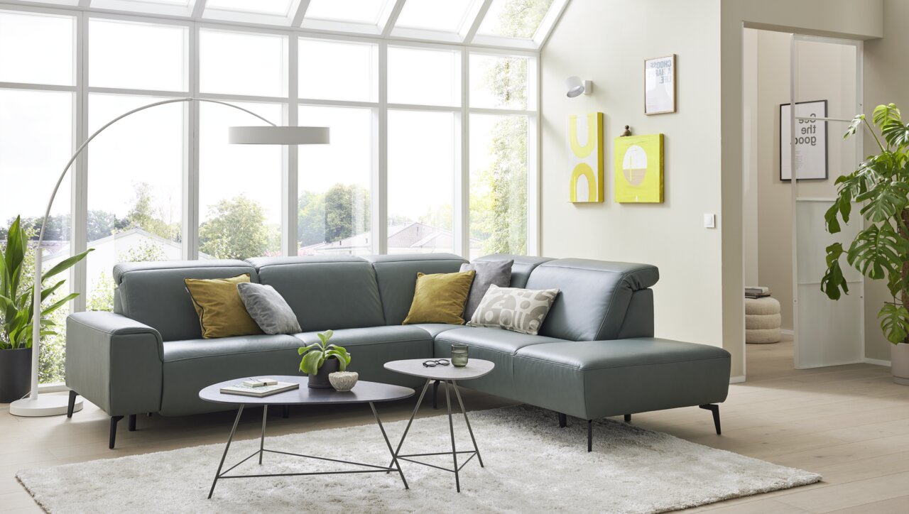 Modernes Ecksofa in einem hellen Wohnzimmer mit großen Fenstern, grauen Polstern und dekorativen Kissen, seitliche Perspektive.