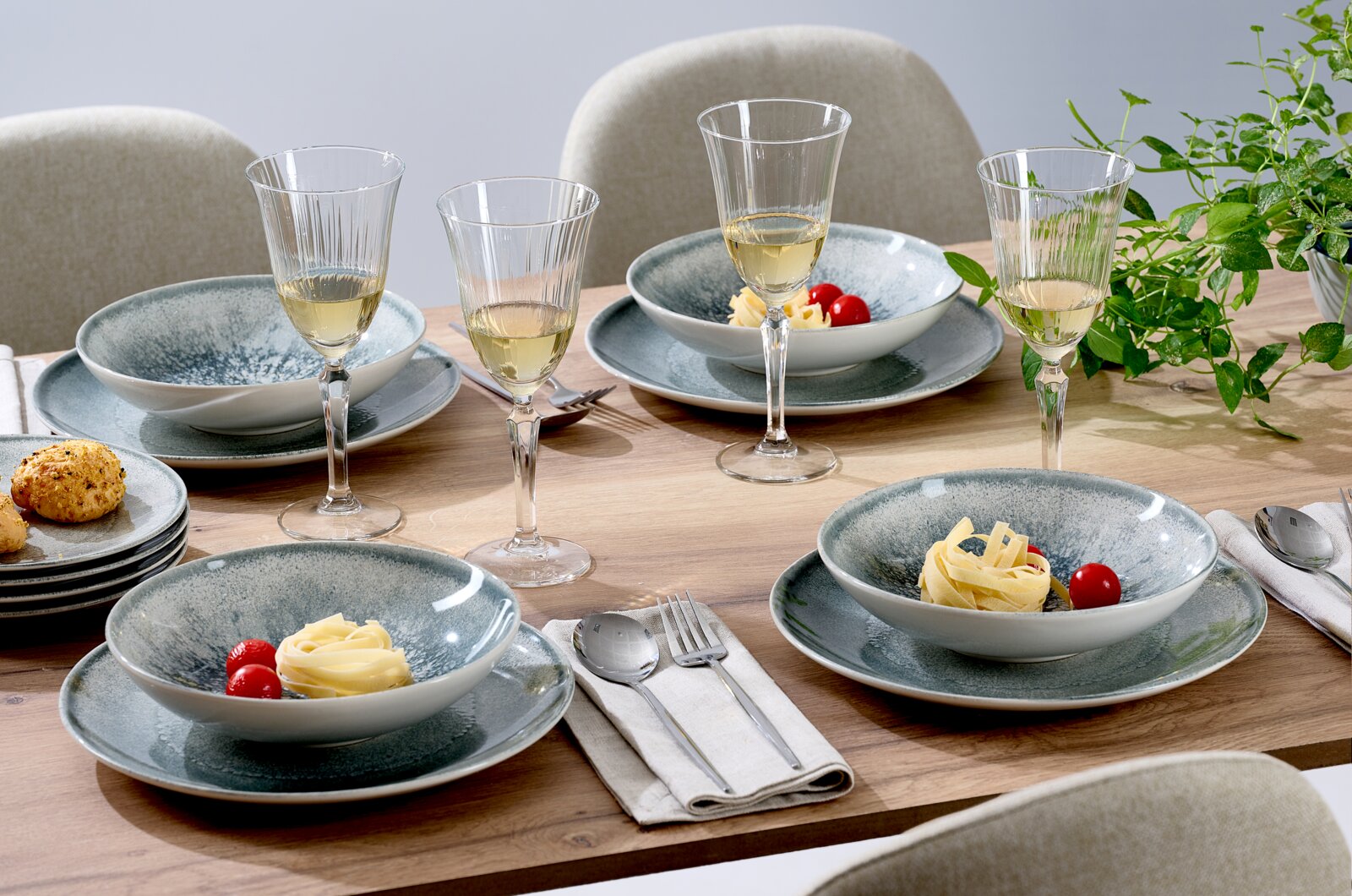 Schuin vooraanzicht van een stijlvol gedekte houten eettafel met blauwgrijs keramisch servies, geribbelde wijnglazen, bestek en groene planten.