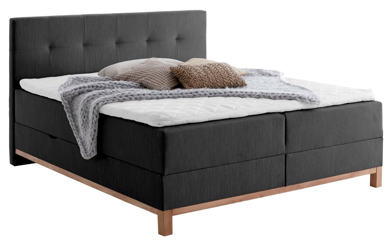 Boxspringbed met donkere bekleding en een hoog hoofdeinde, versierd met kussens en een deken, zijaanzicht