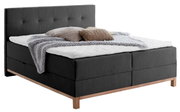 Boxspringbed met donkere bekleding en een hoog hoofdeinde, versierd met kussens en een deken, zijaanzicht