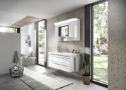 Modern badkamermeubel in een lichte badkamer met grote ramen die uitzicht bieden op het groen. Het badkamermeubel bestaat uit een witte wastafel met twee lades en een verlichte spiegel erboven. Perspectief: zijaanzicht.