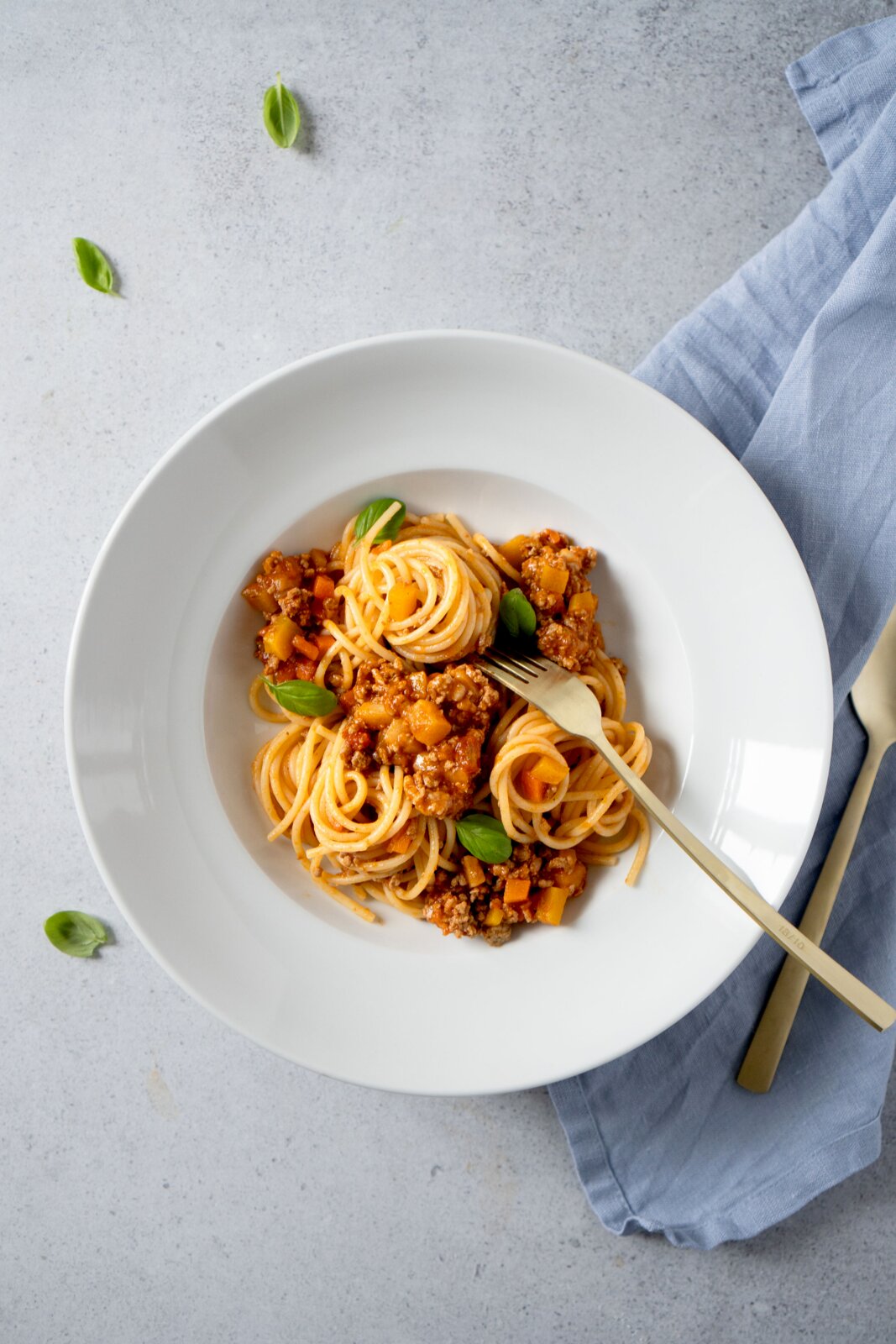 Spaghetti mit Bolognese-Sauce und Basilikum in einem weißen tiefen Teller, daneben goldfarbenes Besteck auf blauem Tuch, Draufsicht (Top-Down)