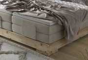 Nahaufnahme eines Boxspringbetts mit grauem Stoffbezug und Holzrahmen, von der Seite betrachtet.