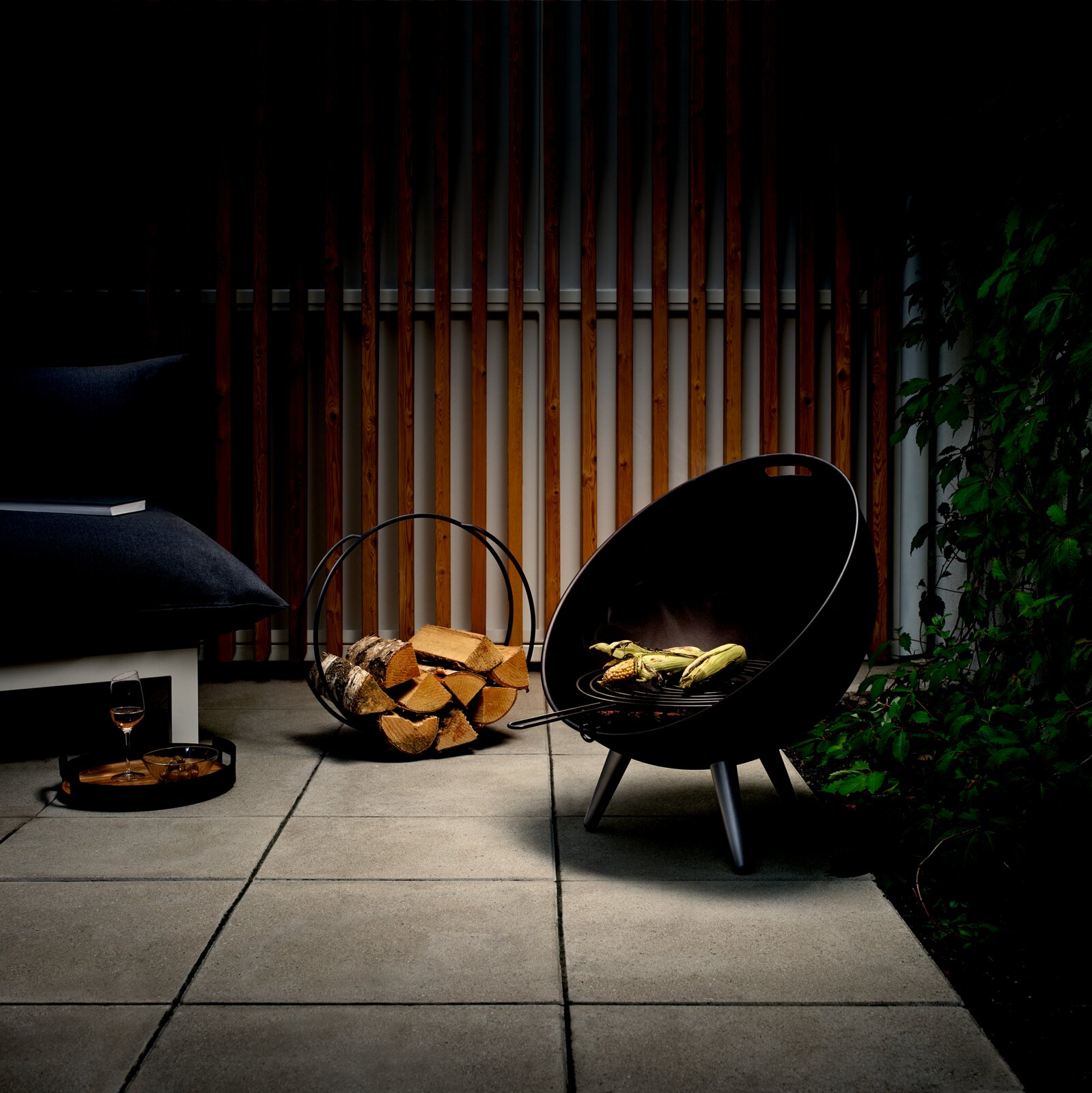 Moderner FireGlobe Grillrost in einem stilvollen Garten, seitliche Perspektive mit brennendem Holz und gegrilltem Gemüse.
