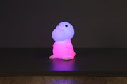 Niedliche Akku-Nachtleuchte in Dinosaurierform, die in verschiedenen Farben leuchtet, frontal auf einem Tisch platziert.