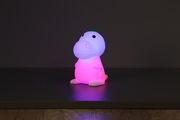 Niedliche Akku-Nachtleuchte in Dinosaurierform, die in verschiedenen Farben leuchtet, frontal auf einem Tisch platziert.