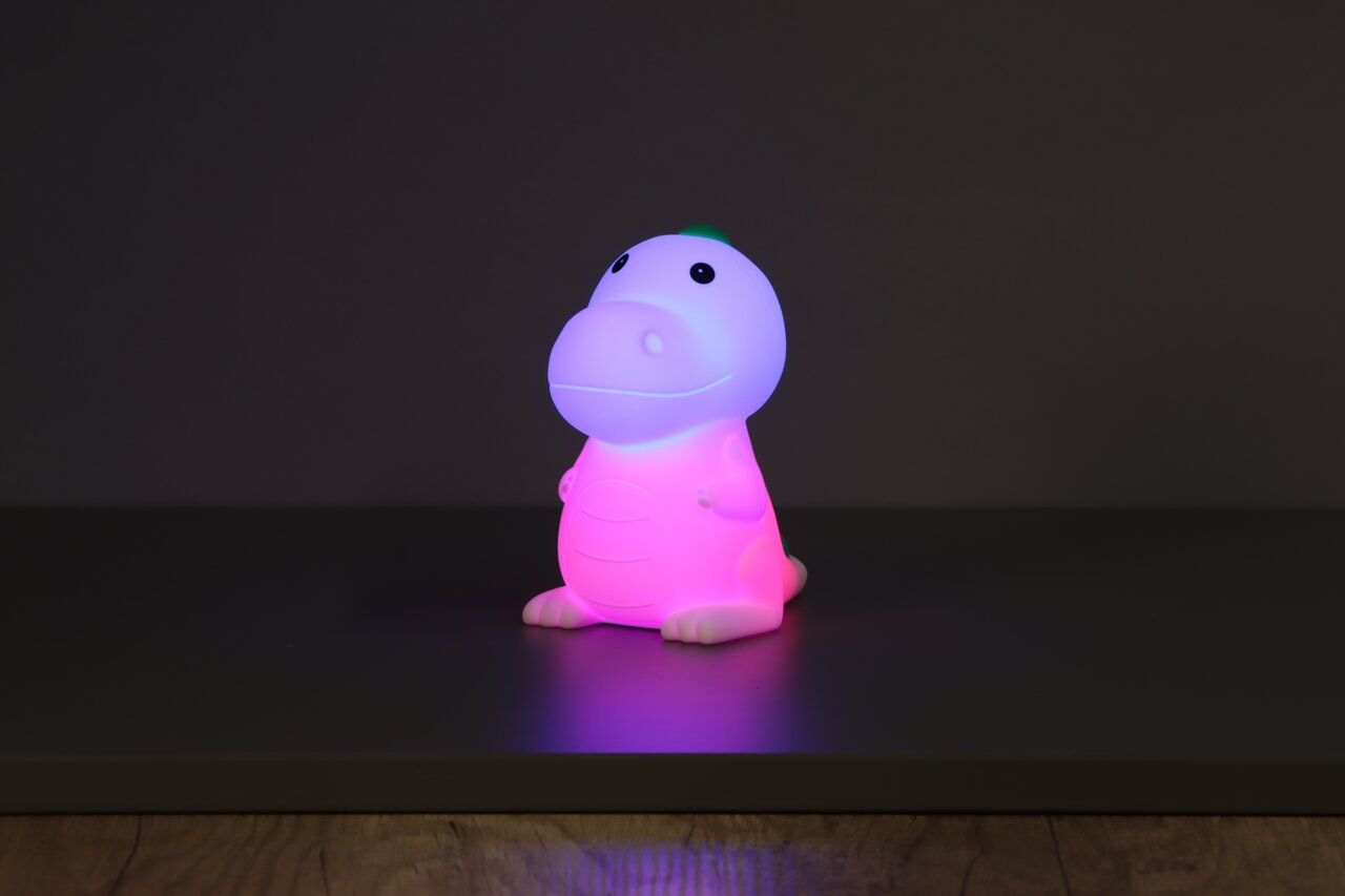 Schattig nachtlampje op batterijen in de vorm van een dinosaurus, dat in verschillende kleuren licht geeft, vooraan op een tafel geplaatst.