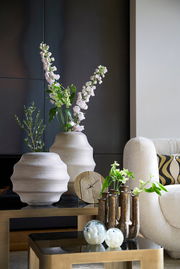 Moderne Wohnzimmerdekoration mit einer goldenen Uhr auf einem Tisch, umgeben von stilvollen Vasen und Pflanzen, seitliche Perspektive.