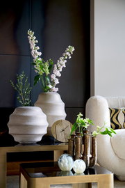 Moderne Wohnzimmerdekoration mit einer goldenen Uhr auf einem Tisch, umgeben von stilvollen Vasen und Pflanzen, seitliche Perspektive.