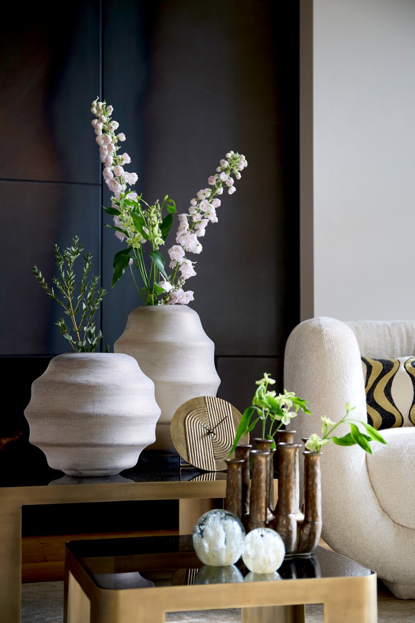 Moderne Wohnzimmerdekoration mit einer goldenen Uhr auf einem Tisch, umgeben von stilvollen Vasen und Pflanzen, seitliche Perspektive.