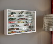 Weiße Wandvitrine mit Glasfront und mehreren Glasböden, gefüllt mit Modellautos und -fahrzeugen, im Raum neben einer gestreiften Stehlampe, perspektivisch in leichter Seitenansicht von vorne