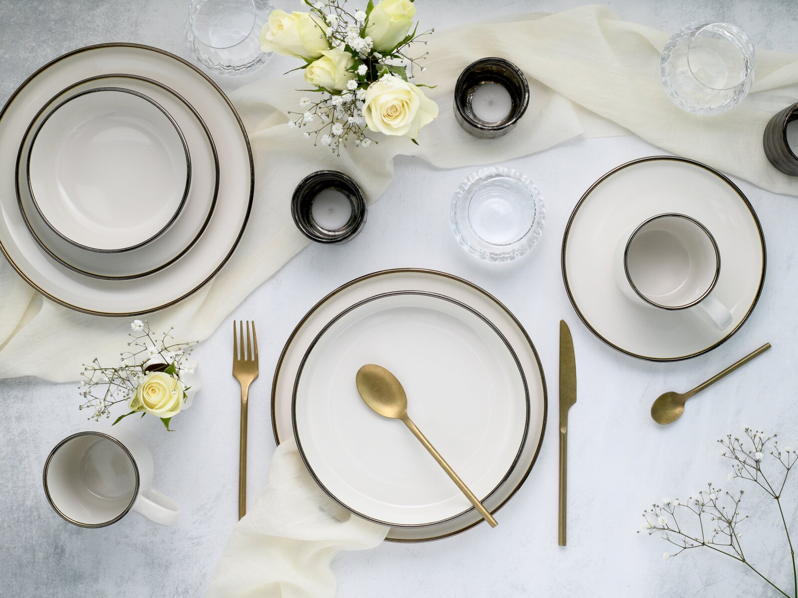 Draufsicht auf ein elegantes weißes Geschirrset mit goldfarbenem Rand (Teller, Schalen und Tasse) auf heller Tischdecke, kombiniert mit goldfarbenem Besteck, Glas-Teelichthaltern und weißen Rosen.