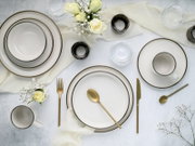 Draufsicht auf ein elegantes weißes Geschirrset mit goldfarbenem Rand (Teller, Schalen und Tasse) auf heller Tischdecke, kombiniert mit goldfarbenem Besteck, Glas-Teelichthaltern und weißen Rosen.