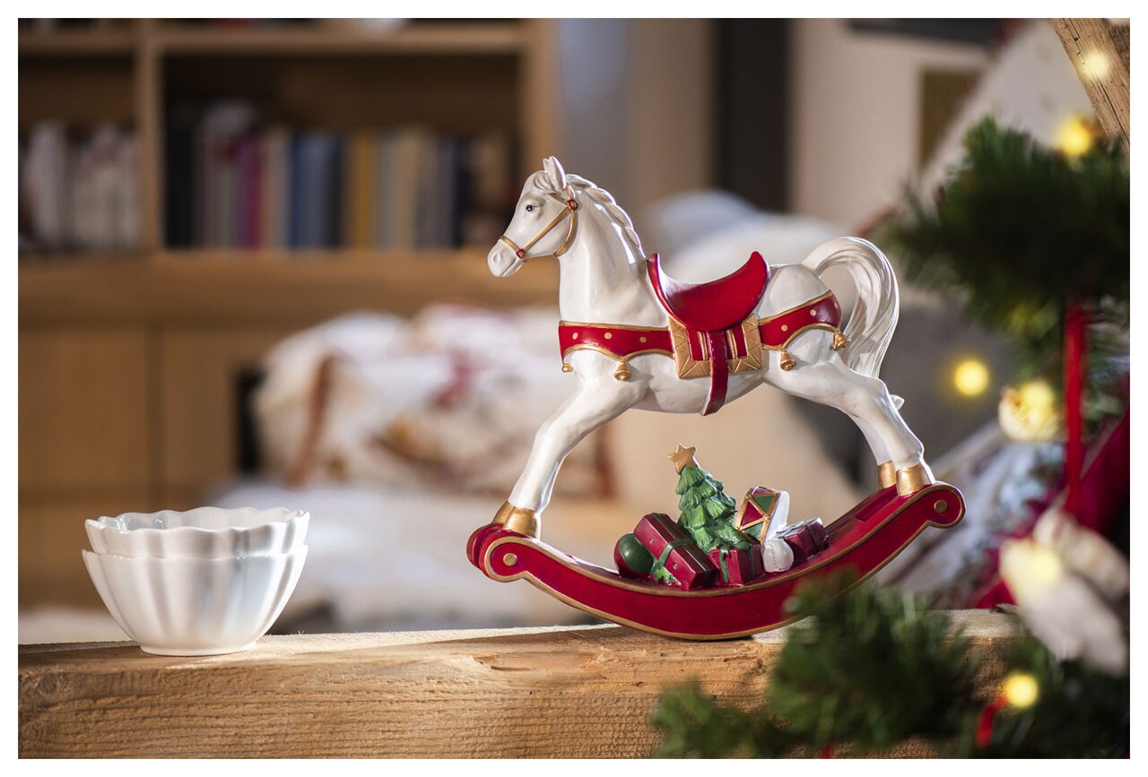 Weißes Schaukelpferd mit rotem Sattel und goldenen Details, seitliche Perspektive, dekoriert mit Geschenken und einem Weihnachtsbaum auf dem Sockel.