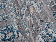Nahaufnahme eines Designer Teppichs mit grauem Hintergrund und blauen, beigen sowie weißen Akzenten. Die Perspektive zeigt die Textur und das Muster des Teppichs in einer seitlichen Ansicht.