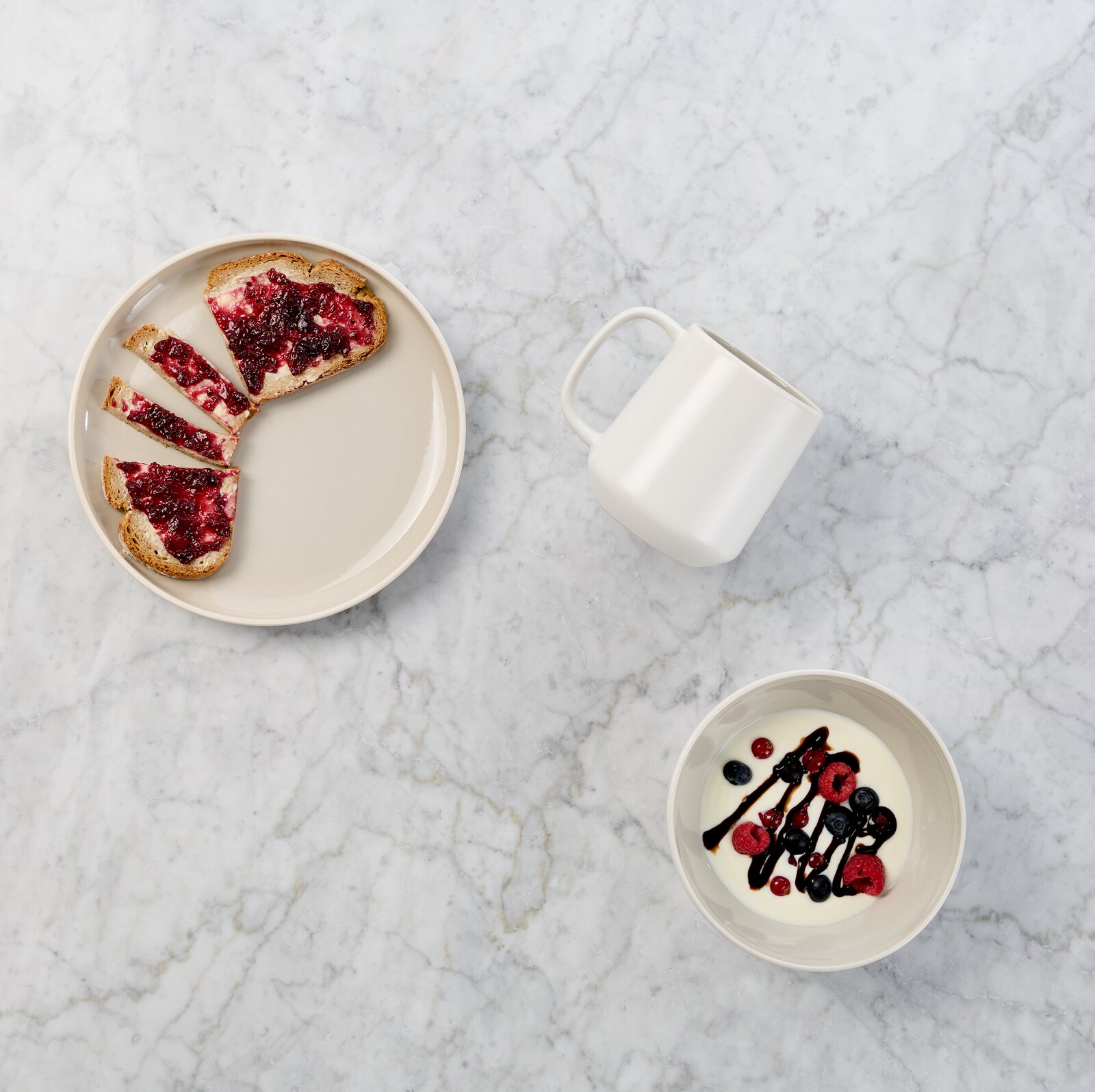 Draufsicht auf ein minimalistisches Frühstücks-Arrangement auf heller Marmorfläche: beiger Teller mit Toast und roter Marmelade, weiße Tasse und Schale mit Joghurt, Beeren und dunkler Sauce.