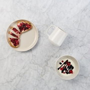 Draufsicht auf ein minimalistisches Frühstücks-Arrangement auf heller Marmorfläche: beiger Teller mit Toast und roter Marmelade, weiße Tasse und Schale mit Joghurt, Beeren und dunkler Sauce.