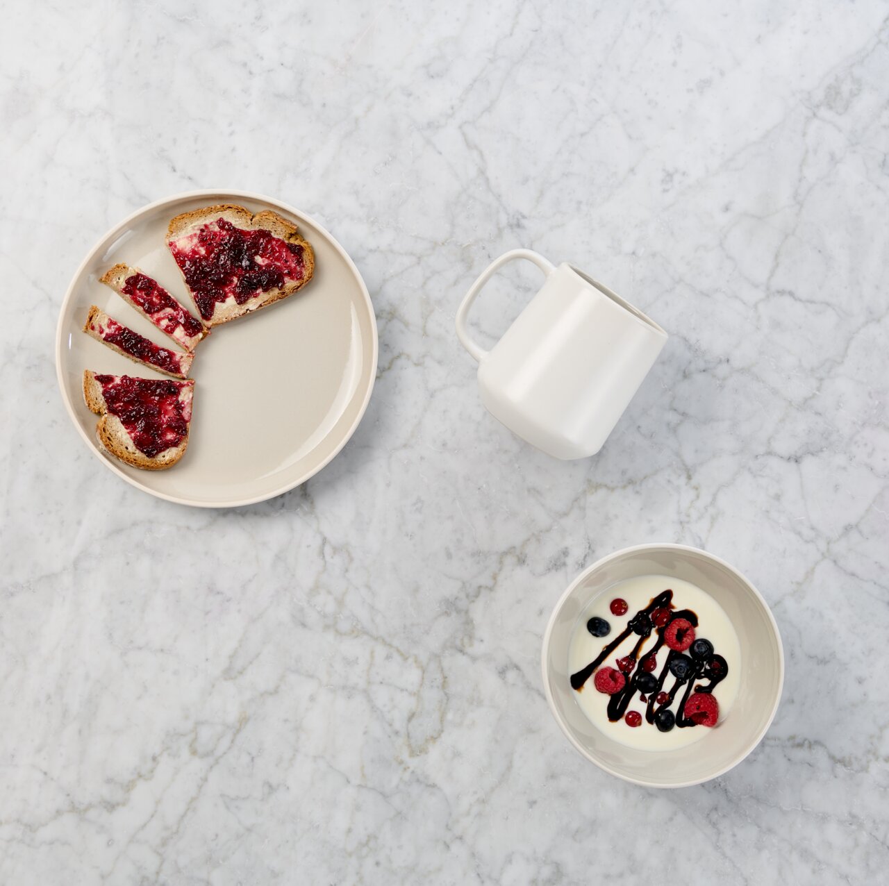 Draufsicht auf ein minimalistisches Frühstücks-Arrangement auf heller Marmorfläche: beiger Teller mit Toast und roter Marmelade, weiße Tasse und Schale mit Joghurt, Beeren und dunkler Sauce.