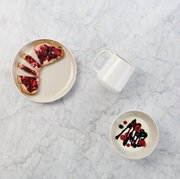Bovenaanzicht van een minimalistisch ontbijt op een licht marmeren oppervlak: een beige bord met toast en rode jam, een wit kopje en een schaaltje met yoghurt, bessen en donkere saus.