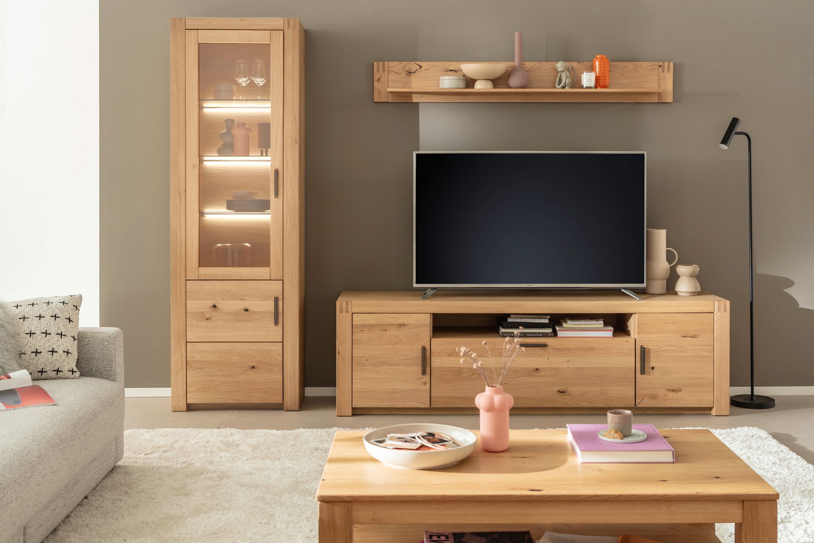 Moderne Wohnzimmeransicht mit einer beleuchteten Vitrine aus Holz, einem TV-Schrank und einem Couchtisch, alles in hellem Holzton. Perspektive von vorne.