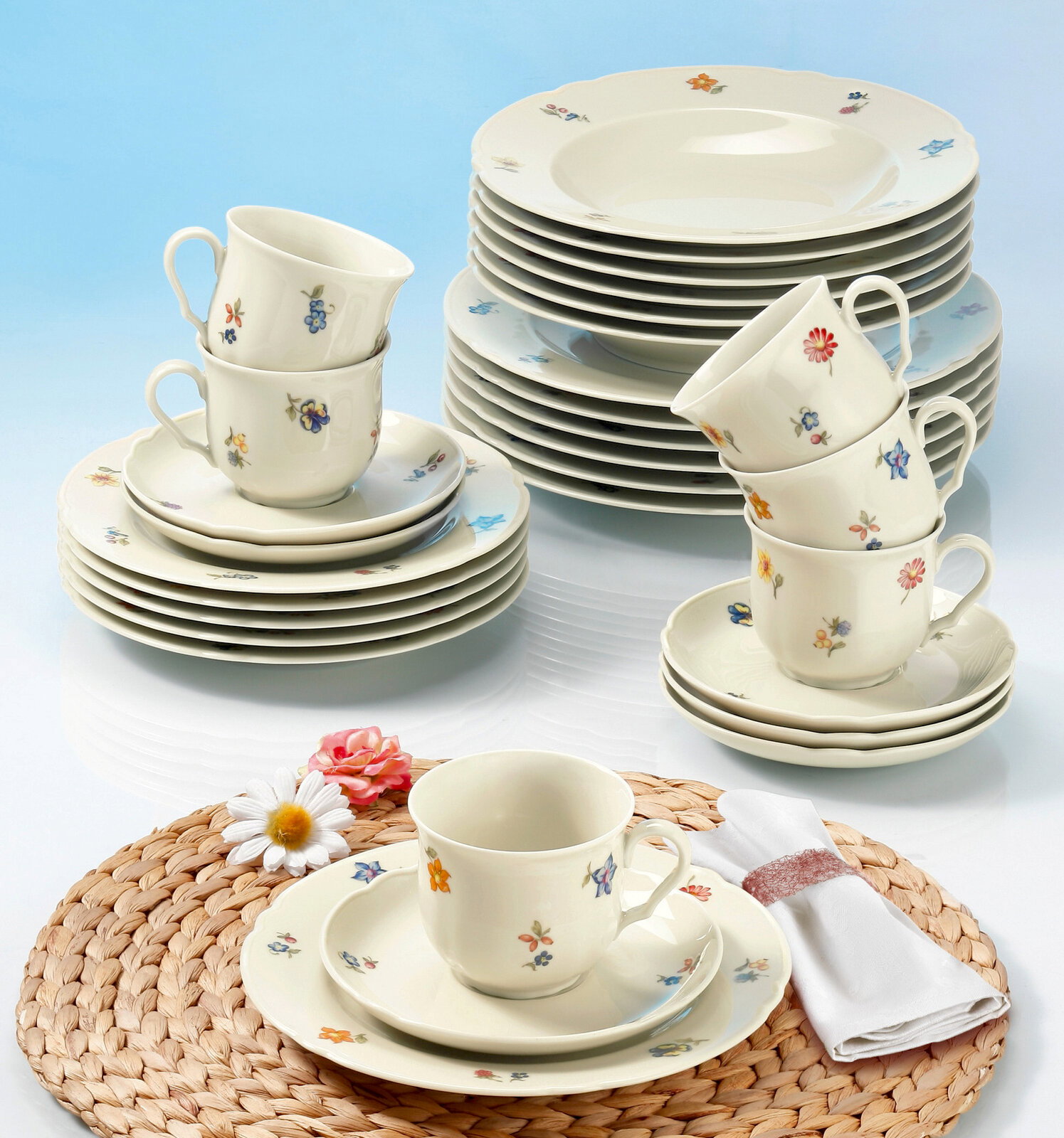 Kaffeeset Marie-Luise 18-tlg. mit floralem Muster, bestehend aus Tassen, Untertassen und Tellern, auf einem Tisch mit geflochtener Unterlage und Blumen, aus der Vogelperspektive fotografiert.