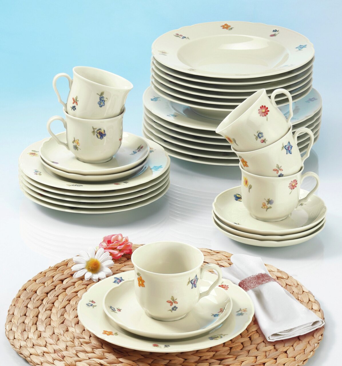 Kaffeeset Marie-Luise 18-tlg. mit floralem Muster, bestehend aus Tassen, Untertassen und Tellern, auf einem Tisch mit geflochtener Unterlage und Blumen, aus der Vogelperspektive fotografiert.