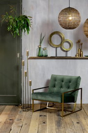 Ein stilvolles Wohnzimmer mit einem grünen Sessel aus Samt und goldenen Metallakzenten, umgeben von modernen Dekorationen wie hängenden Lampen und Pflanzen. Perspektive: Frontalansicht.