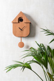 Moderne Wanduhr aus Holz in Form eines Vogelhauses mit Pendel, an einer hellen Wand montiert, mit grüner Pflanze im Vordergrund, seitliche Perspektive.