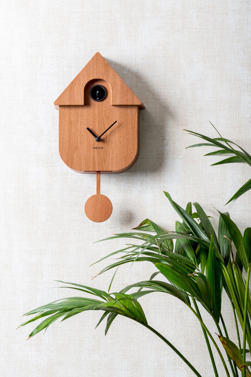 KARLSSON Wandklok CUCKOO Moderne houten wandklok in de vorm van een vogelhuisje met slinger, bevestigd aan een lichte muur, met een groene plant op de voorgrond, zijaanzicht.