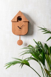 Moderne Wanduhr aus Holz in Form eines Vogelhauses mit Pendel, an einer hellen Wand montiert, mit grüner Pflanze im Vordergrund, seitliche Perspektive.