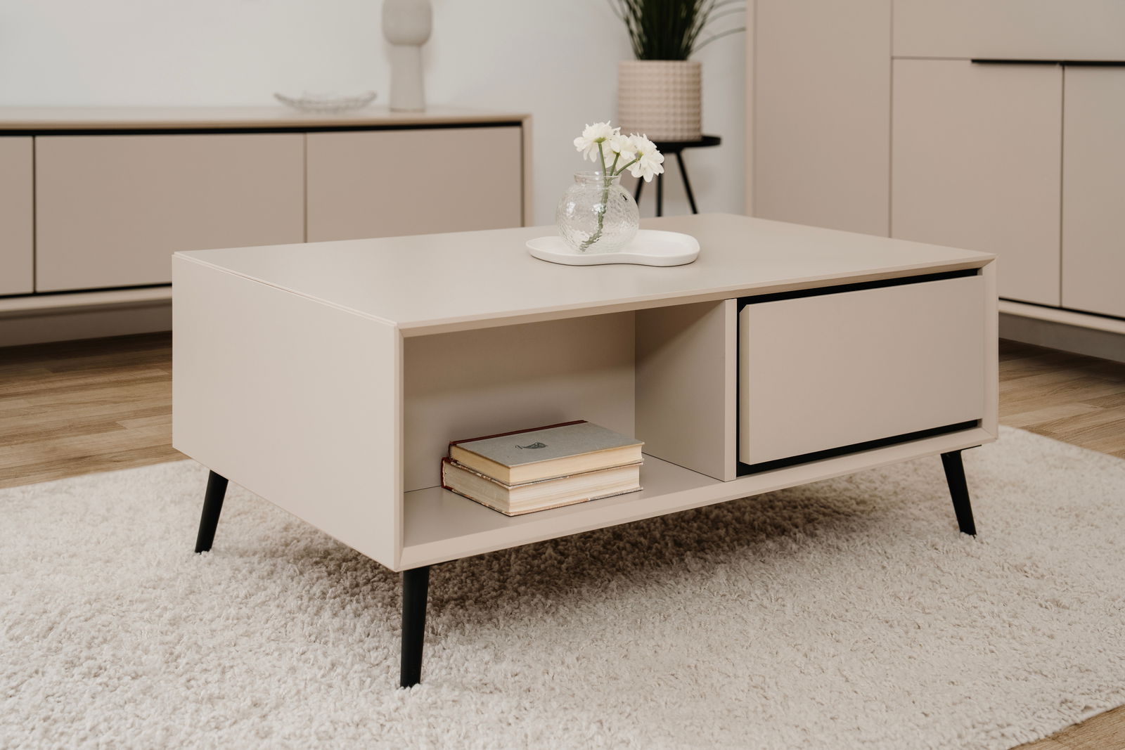 Beige Couchtisch mit schwarzem Gestell, dekoriert mit einer Vase und Büchern, aus seitlicher Perspektive fotografiert.