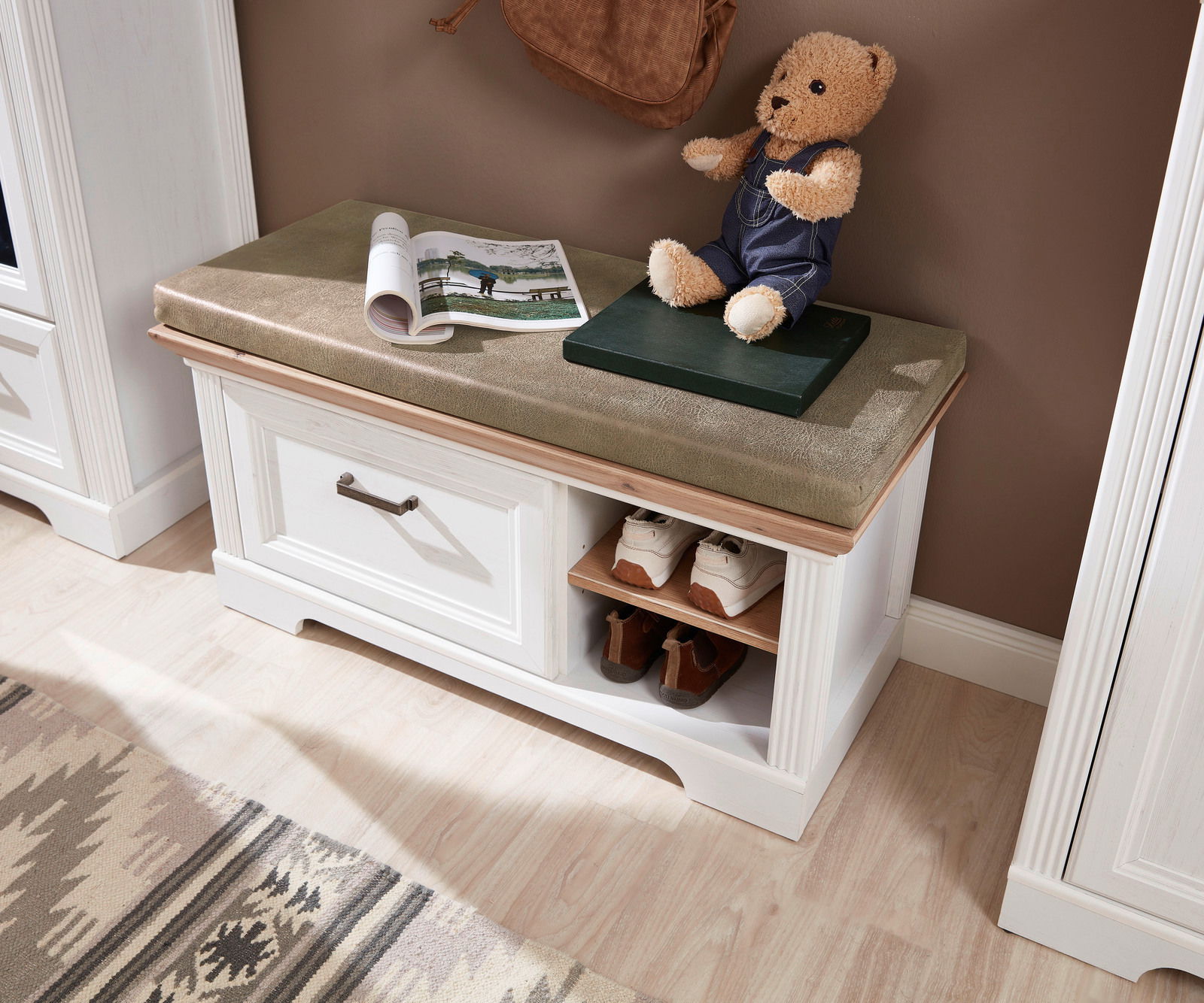 Garderobenbank mit weißem Holzrahmen, grünem Sitzpolster, Stauraum für Schuhe und dekorativem Teddybär, aus der Vogelperspektive.