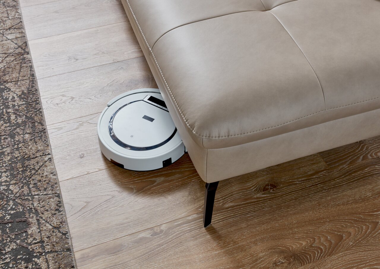 Een robotstofzuiger maakt de vloer schoon onder een beige hoekbank, gezien vanuit vogelperspectief.
