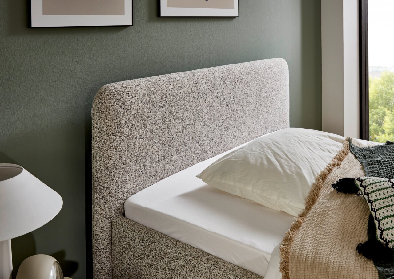 Seitliche Perspektive eines Polsterbetts mit grauem Kopfteil, weißem Bettlaken und dekorativen Kissen in einem modern eingerichteten Raum.